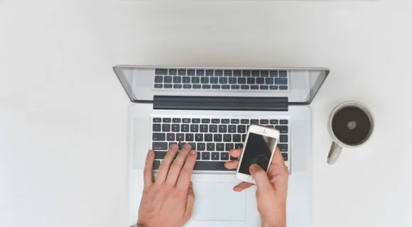 A laptop on a white table with a cup of coffee and a pair of hands holding a phone
