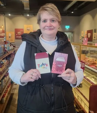 A woman dressed in beige and black holds up two Costa coffee gift cards